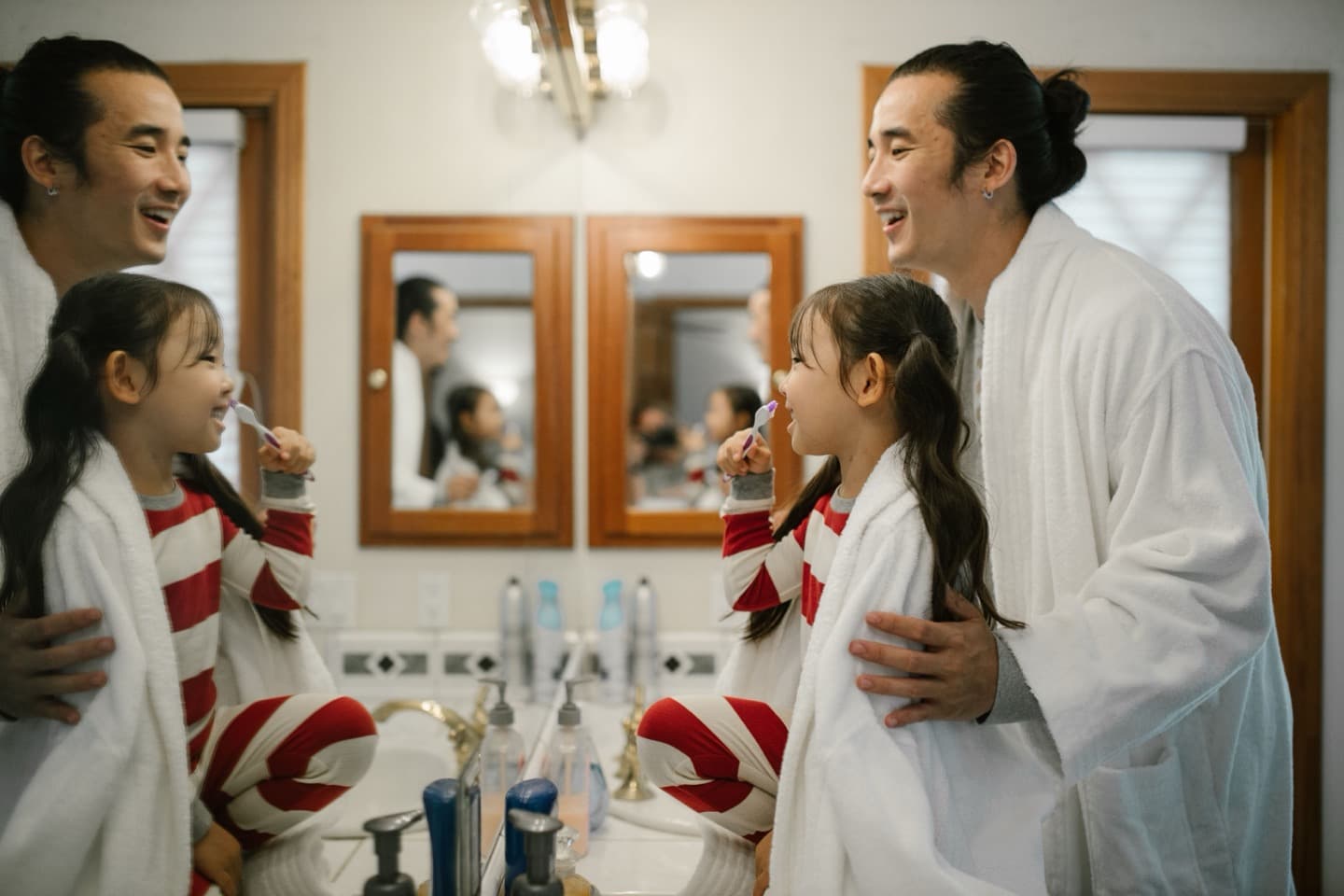 A father helping his daughter brush her teeth in a bright bathroom, both smiling